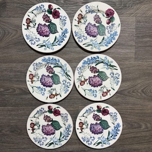 May include: A set of six round white ceramic plates, each featuring an identical botanical illustration of purple hydrangeas, dark red blossoms, and light blue wildflowers with green leaves arranged in a circular pattern around the center.