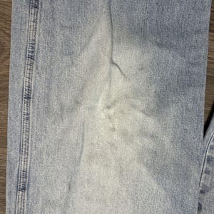 May include: Close-up of light blue denim jeans. The jeans show signs of wear and fading, with lighter patches and subtle creases. The stitching along the seam is visible, adding detail to the fabric.