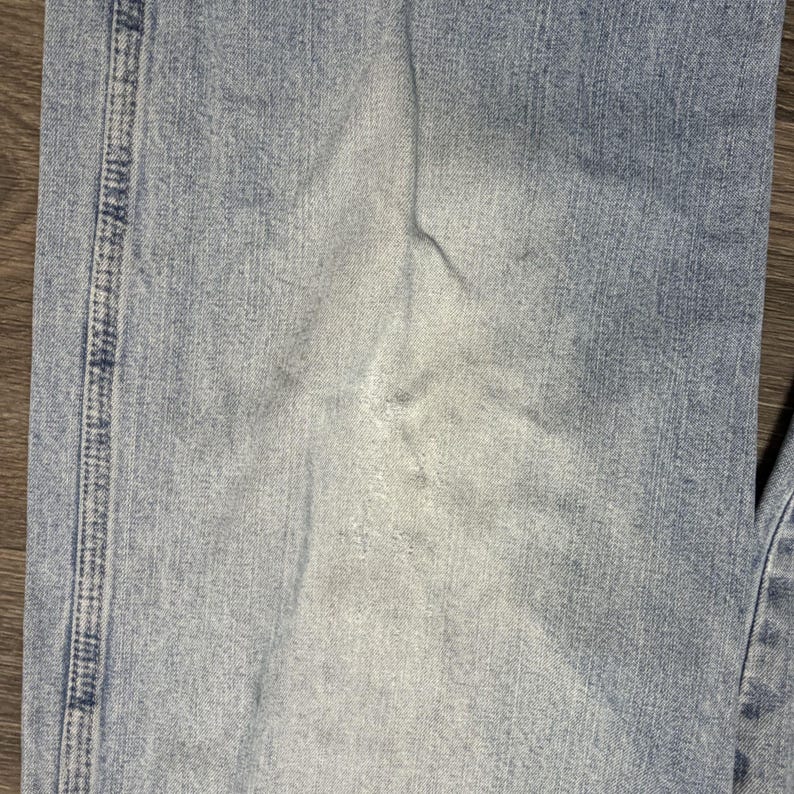 May include: Close-up of light blue denim jeans. The jeans show signs of wear and fading, with lighter patches and subtle creases. The stitching along the seam is visible, adding detail to the fabric.