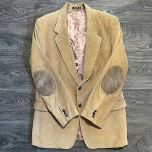 May include: Tan corduroy blazer with suede elbow patches. The jacket has a classic design with a notched lapel, three-button closure, and buttoned cuffs. The interior lining is a light pink color.