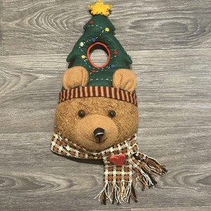 May include: A plush bear head ornament featuring a dark green felt Christmas tree top with a yellow star, a brown striped hat band, and a brown and white checkered scarf with a small red heart accent. The bear has dark eyes and a black nose.