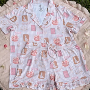 Puede incluir: Un conjunto de pijama rosa y blanco con una parte superior de manga corta y pantalones cortos. La parte superior tiene un cuello abotonado y los pantalones cortos tienen una cintura con cord&oacute;n. La tela presenta un patr&oacute;n repetido de una novia y un novio, corazones y flores.