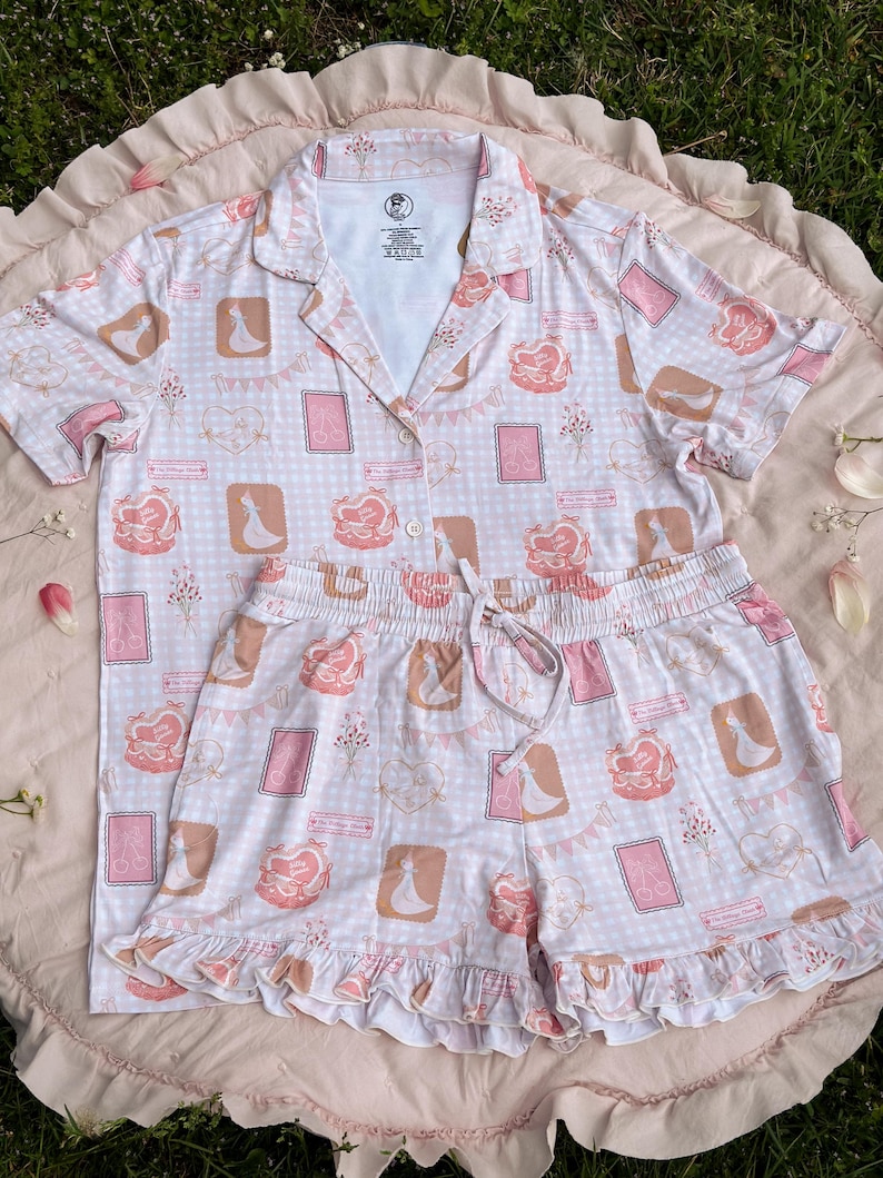 Puede incluir: Un conjunto de pijama rosa y blanco con una parte superior de manga corta y pantalones cortos. La parte superior tiene un cuello abotonado y los pantalones cortos tienen una cintura con cord&oacute;n. La tela presenta un patr&oacute;n repetido de una novia y un novio, corazones y flores.