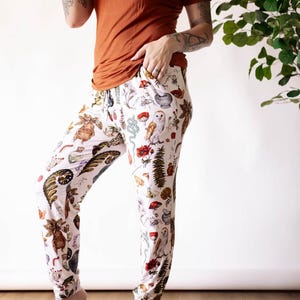 May include: White pants with a colorful print of various botanical illustrations, including flowers, leaves, and insects. The pants have a drawstring waistband and a relaxed fit.
