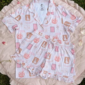 Puede incluir: Un conjunto de pijama rosa y blanco con estampado de cuadros vichy y estampado de cartas de juego. El conjunto incluye una camisa de manga corta con cuello y un pantal&oacute;n corto con ribete de volantes.
