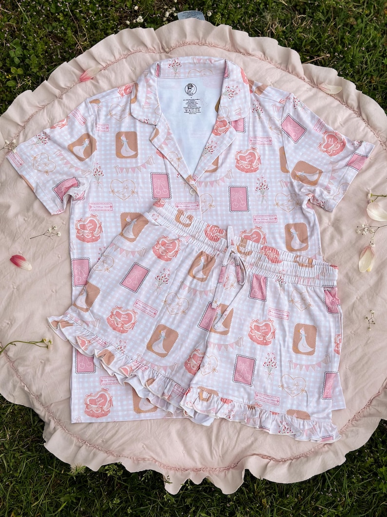 Puede incluir: Un conjunto de pijama rosa y blanco con estampado de cuadros vichy y estampado de cartas de juego. El conjunto incluye una camisa de manga corta con cuello y un pantal&oacute;n corto con ribete de volantes.