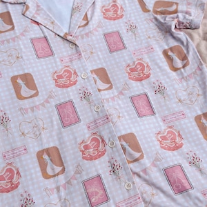 Puede incluir: Una camisa de botones rosa y blanca con estampado de cuadros vichy. La camisa presenta un patr&oacute;n repetitivo de gansos, corazones y flores. El texto "The Village Goose" se repite en todo el patr&oacute;n.