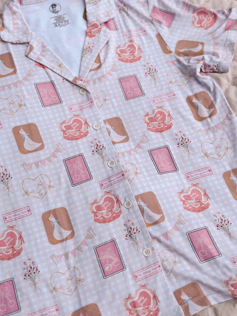 Puede incluir: Una camisa de botones rosa y blanca con estampado de cuadros vichy. La camisa presenta un patr&oacute;n repetitivo de gansos, corazones y flores. El texto "The Village Goose" se repite en todo el patr&oacute;n.