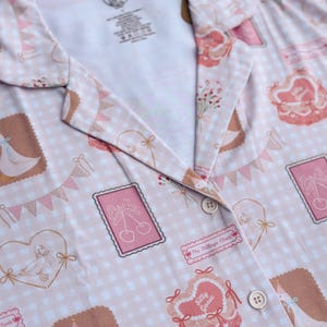 Puede incluir: Una camisa de botones rosa y blanca con estampado de cuadros vichy y una variedad de ilustraciones lindas, que incluyen gansos, corazones, cerezas y flores. La camisa tiene cuello y dos botones. El texto "The Village Cloth" y "Silly Goose" est&aacute; impreso en la camisa.