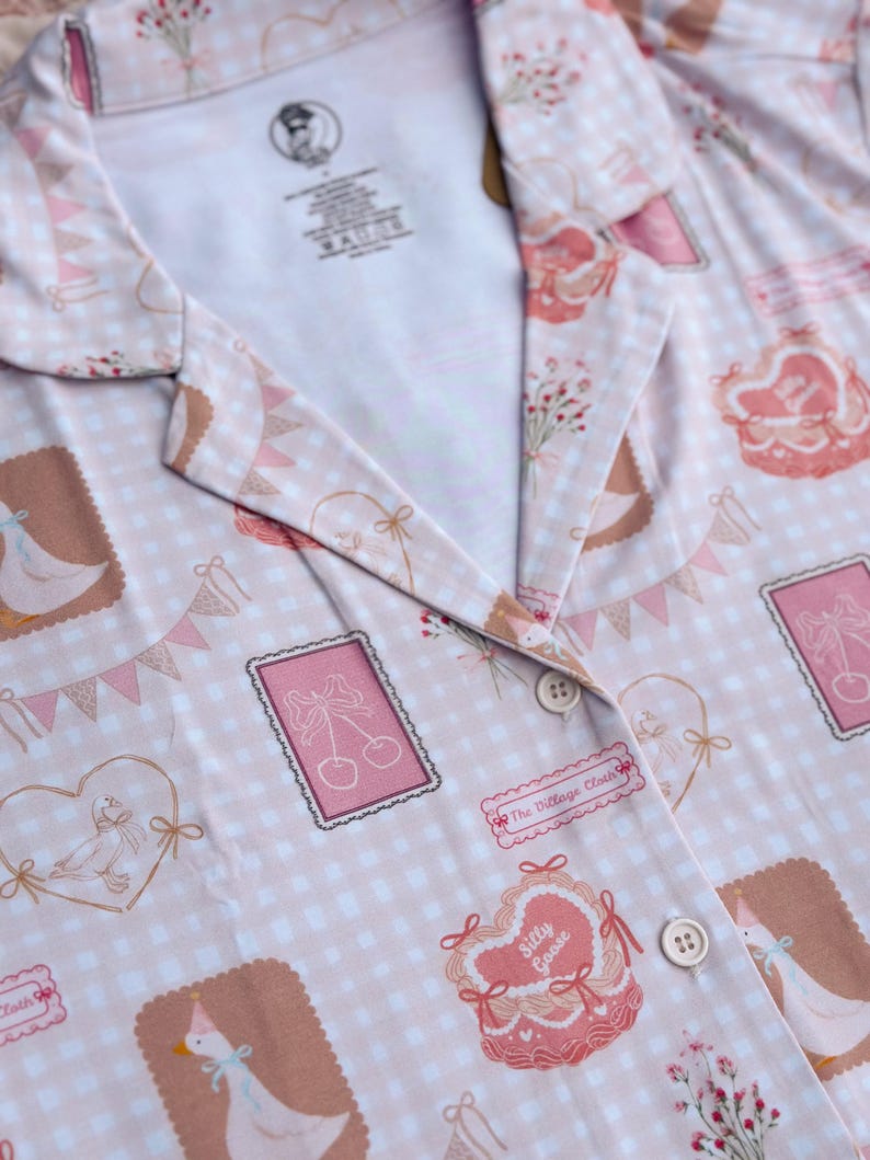 Puede incluir: Una camisa de botones rosa y blanca con estampado de cuadros vichy y una variedad de ilustraciones lindas, que incluyen gansos, corazones, cerezas y flores. La camisa tiene cuello y dos botones. El texto "The Village Cloth" y "Silly Goose" est&aacute; impreso en la camisa.