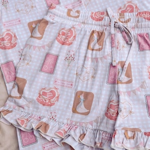 Puede incluir: Conjunto de pijama rosa y blanco con estampado de cuadros vichy y estampado de ganso. El conjunto incluye una parte superior de manga corta con cierre de botones en la parte delantera y un pantal&oacute;n corto con dobladillo con volantes.