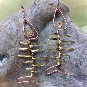 May include: A pair of handmade earrings in the shape of a fish skeleton. The earrings are crafted from copper and gold-colored wire, with a small, yellow bead accent in the top triangle. The earrings are displayed on a piece of driftwood.