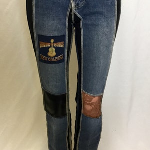 May include: Blue denim jeans with black side panels and patches. One patch reads "HOUSE OF BLUES NEW ORLEANS" with a guitar graphic. Features a black leather patch and a brown lace patch. The jeans have a button closure.