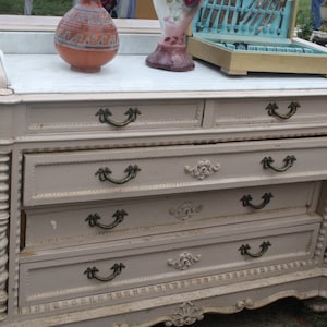 May include: A vintage, light pink dresser with five drawers and ornate details. The dresser has a marble top and decorative carved details on the front and sides. The drawers have antique brass handles.