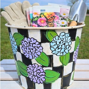 May include: A decorative garden bucket with a black and white checkered pattern and painted blue and purple flowers. The bucket contains gardening gloves, seed packets, and a small trowel. The bucket is sitting on a white surface.