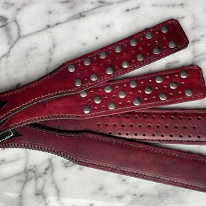 Pode incluir: Quatro tiras de couro bordô com rebites prateados e costuras decorativas. Uma tira apresenta um design em forma de coração com couro preto e costuras brancas. As tiras estão dispostas sobre uma superfície de mármore.