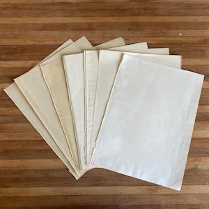 May include: A collection of several rectangular sheets of paper arranged in a fan shape. The papers vary in colour from off-white to light cream and tan, featuring deckled edges on the sides and a smooth, matte texture throughout.