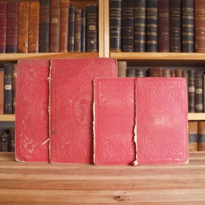 May include: Four red hardcover books with ornate designs on the covers. The books are arranged in a row on a wooden surface.