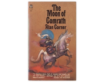 May include: A vintage paperback book cover with a fantasy illustration of a knight riding a white horse. The book title is "The Moon of Comrath" by Alan Garner. The cover is brown with a blue and white sky in the background.