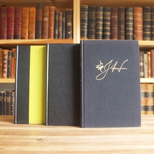 May include: Three hardcover books with black covers. The book on the left has a yellow spine. The book on the right has the initials "J.H." in gold lettering on the front cover.