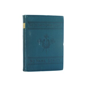 May include: A blue hardcover book with the title "The Colonel's Opera Cloak" and the text "No Name Series" on the cover. The book features a horseshoe and clover design.