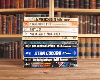 May include: A collection of vintage science fiction books by Keith Laumer, such as "Star Colony" and "The Shape Changer." The books display various colours and designs, with visible titles and author names. A wooden bookshelf filled with books forms the background.