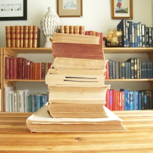 May include: A stack of vintage hardback books with brown and cream coloured covers on a wooden table. The books are arranged in a bookshelf behind the stack.