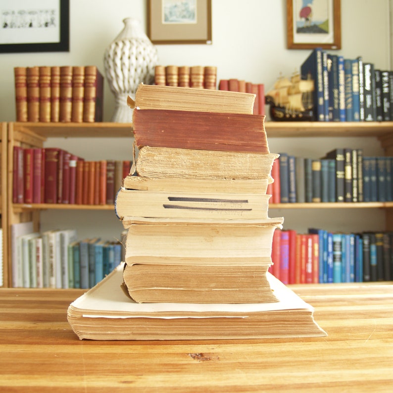 May include: A stack of vintage hardback books with brown and cream coloured covers on a wooden table. The books are arranged in a bookshelf behind the stack.