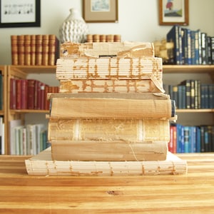 May include: A stack of vintage books with worn edges and yellowed pages. The books are arranged on a wooden surface with a bookshelf in the background.