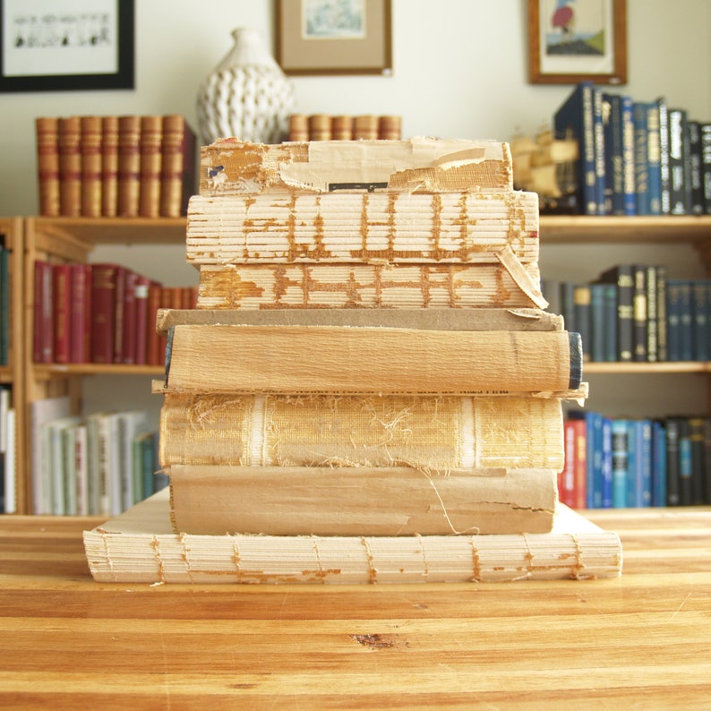 May include: A stack of vintage books with worn edges and yellowed pages. The books are arranged on a wooden surface with a bookshelf in the background.