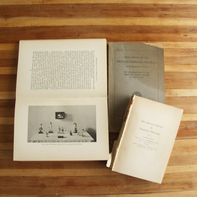 May include: Three vintage books on a wooden surface. The book on the left has a black and white photo of a display of small figurines. The book in the middle has the title "Final Report of the Provost Marshal General" and the book on the right has the title "The Oxford Library of Practical Theology".