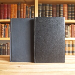 May include: Two black hardcover books with blank spines, sitting on a wooden surface. The books are in front of a bookshelf filled with books.