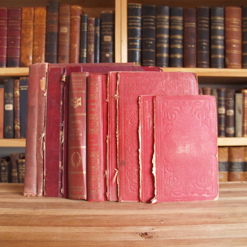 May include: A stack of red hardcover books with gold lettering on the spines. The books are arranged on a wooden surface.