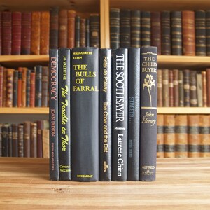 May include: A row of ten black hardcover books with white text titles on the spines. The books are arranged on a wooden table. The titles include "Democracy", "The Trouble in Thor", "The Bulls of Parral", "The Crow and the Cat", "The Soothsayer", "Streets", and "The Child Buyer".