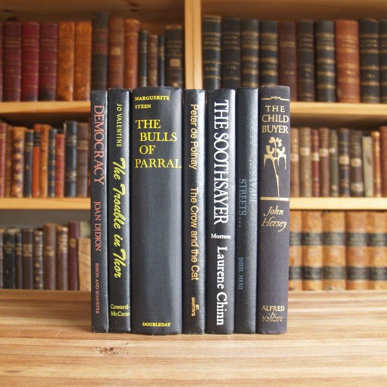 May include: A row of ten black hardcover books with white text titles on the spines. The books are arranged on a wooden table. The titles include "Democracy", "The Trouble in Thor", "The Bulls of Parral", "The Crow and the Cat", "The Soothsayer", "Streets", and "The Child Buyer".