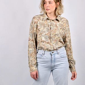 Könnte beinhalten: Ein Hemd mit floralem Muster in Beige-, Braun- und Rosatönen. Das langärmlige Hemd wird mit hellblauen Jeans kombiniert. Das Hemd hat einen klassischen Kragen und geknöpfte Manschetten.