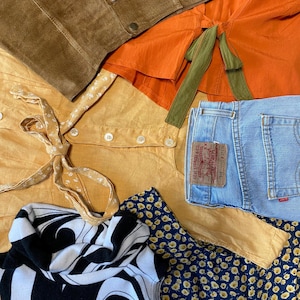 Könnte beinhalten: Eine flache Anordnung verschiedener Kleidungsstücke, darunter eine braune Wildlederjacke, ein senfgelbes Hemd mit Knöpfen, ein schwarz-weiß gestreiftes Hemd, eine blaue Jeans mit einem Levi's-Etikett, ein Rock mit Blumenmuster und ein orangefarbenes Oberteil mit einem grünen Band.