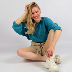 Op de afbeelding: Een turquoise gebreide trui met korte mouwen wordt gecombineerd met een kaki short en witte sneakers. De persoon zit op een witte achtergrond, lacht en raakt haar haar aan.