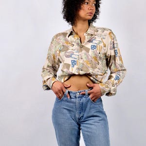 Op de afbeelding: Een beige overhemd met een kleurrijk patroon met vlaggen en andere ontwerpen. Het overhemd heeft lange mouwen en wordt gecombineerd met lichtblauwe jeans. De outfit is casual en vintage-geïnspireerd.