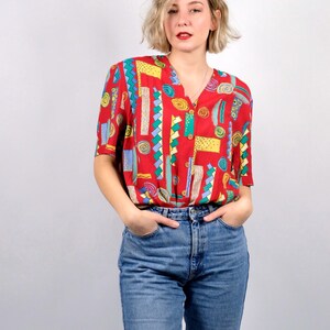 Op de afbeelding: Een rode blouse met korte mouwen en een kleurrijk geometrisch patroon van blauwe, gele en groene vormen. De blouse wordt gecombineerd met een blauwe jeans. De top heeft een V-hals en is aan de voorkant dichtgeknoopt.