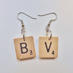 May include: A pair of earrings made from wooden Scrabble tiles. The left earring has the letter "B" with the number "3" on it. The right earring has the letter "V" with the number "4" on it.