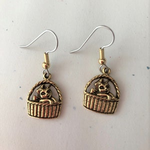 May include: A pair of gold-colored cat earrings. Each earring features a small cat figure sitting inside a woven basket. The earrings have silver-colored ear wires and gold-colored accents.