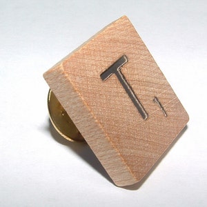 May include: A wooden letter tile with the letter "T" printed on it. The tile is attached to a gold pin.