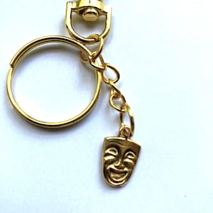 May include: Gold-colored keychain with a lobster clasp, split ring, chain, and a small charm shaped like a smiling theater mask. The keychain is made of shiny metal and is set against a white background.