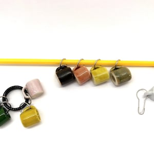 May include: A set of colorful ceramic mug stitch markers, with a yellow knitting needle and a white ceramic mug stitch marker. The stitch markers are attached to the knitting needle with metal rings. The stitch markers are numbered 1 through 7.