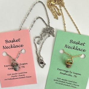 May include: Two silver and gold necklaces with a small basket charm. The necklaces are on pink and green cards with text that reads "Basket Necklace" and "Earrings by Susan Nashville, TN" and "USA-made Lead-free Pewter Charm on Palladium-plated Chain" and "USA-made Lead-free Gold-plated Pewter Charm on Gold-plated Chain".