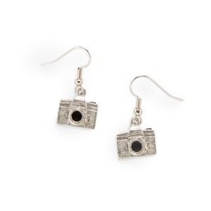 Puede incluir: Pendientes de cámara de plata colgantes. Los pendientes son de plata y tienen un pequeño dije de cámara.