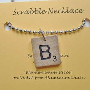 May include: A silver chain necklace with a wooden Scrabble tile pendant featuring the letter "B" and the number "3". The tile is light brown with black lettering. The necklace is on a yellow background with the text "Scrabble Necklace" and "Wooden Game Piece on Nickel-free Aluminum Chain".