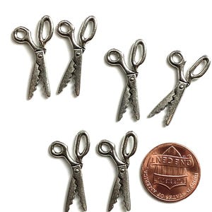 May include: A collection of small, silver-colored scissors charms with a decorative edge. One U.S. penny is included for size comparison. The scissors are arranged on a white background.