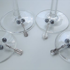 May include: Four wine glass charms with silver musical instrument charms and gray and white speckled beads. The charms include a guitar, a violin, a banjo, and a bass.
